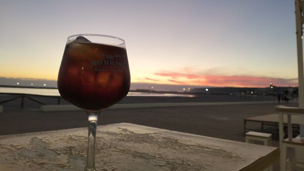 Un verre de vin rouge sur une table, avec vue sur le coucher de soleil sur l’eau.