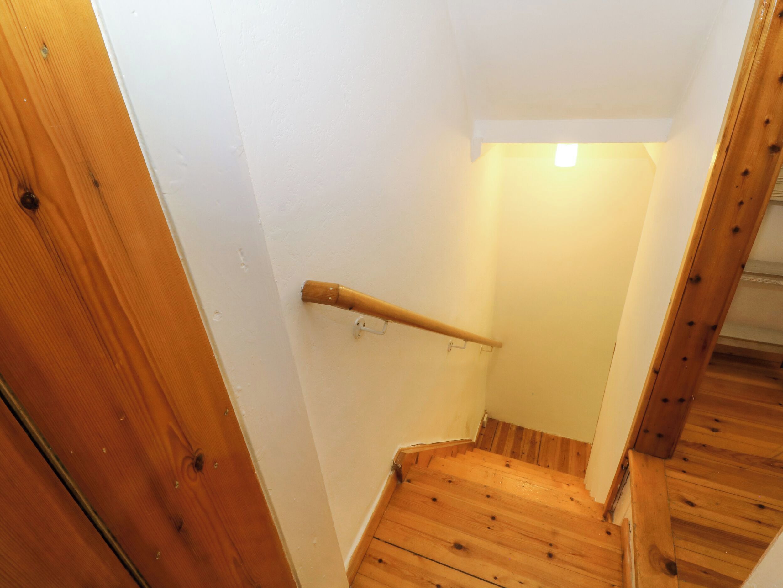A wooden staircase with a handrail, white walls, and a small light at the top.