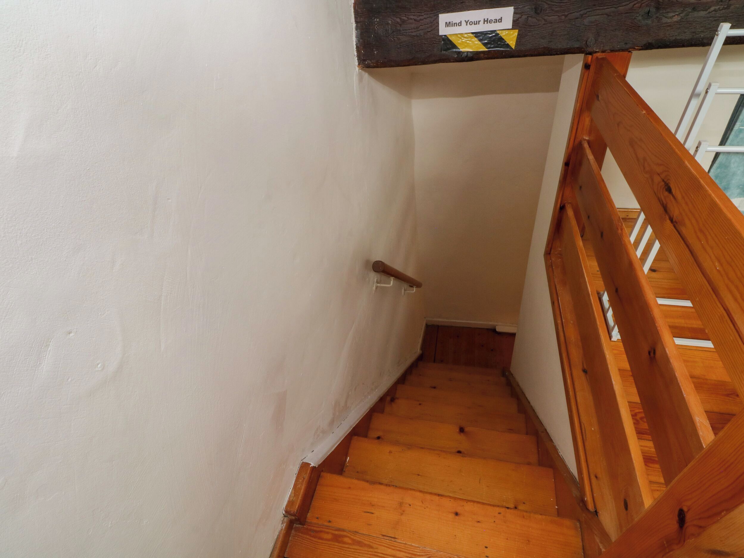 Een houten trap met een leuning, witte muren en een bordje met de tekst 'Let op uw hoofd'.