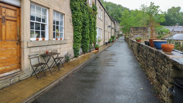 Une rue étroite bordée de maisons mitoyennes, chacune ayant une porte et des fenêtres en bois. Des plantes en pot sont posées sur les rebords de fenêtres, et une petite table avec des chaises se trouve devant l’une des maisons.