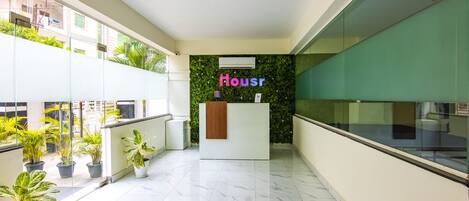 A modern hotel lobby with a reception desk, potted plants, and a green wall with the word "Housr" on it.