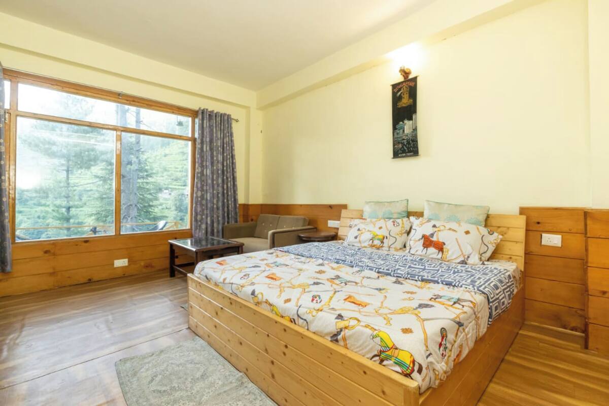 A bedroom with a large bed, a wooden headboard, a desk, and a chair. There is a window with curtains, a poster on the wall, and a view of trees outside.