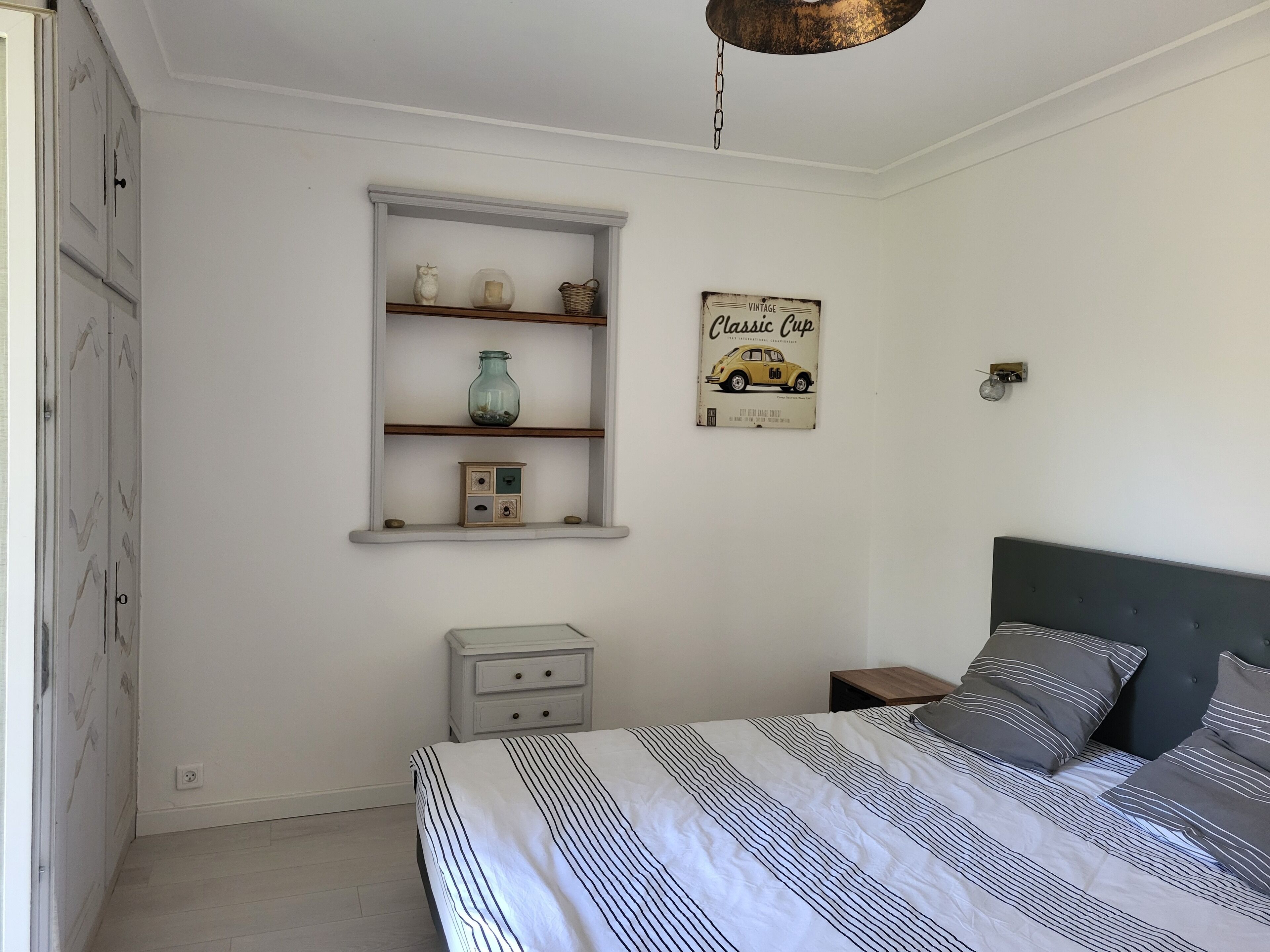 Un dormitorio con cama, mesita de noche, cómoda, estante con objetos decorativos y un cartel de coche antiguo.