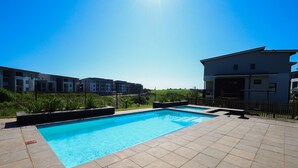 Una piscina con piso de baldosas, rodeada por una valla, y edificios al fondo.
