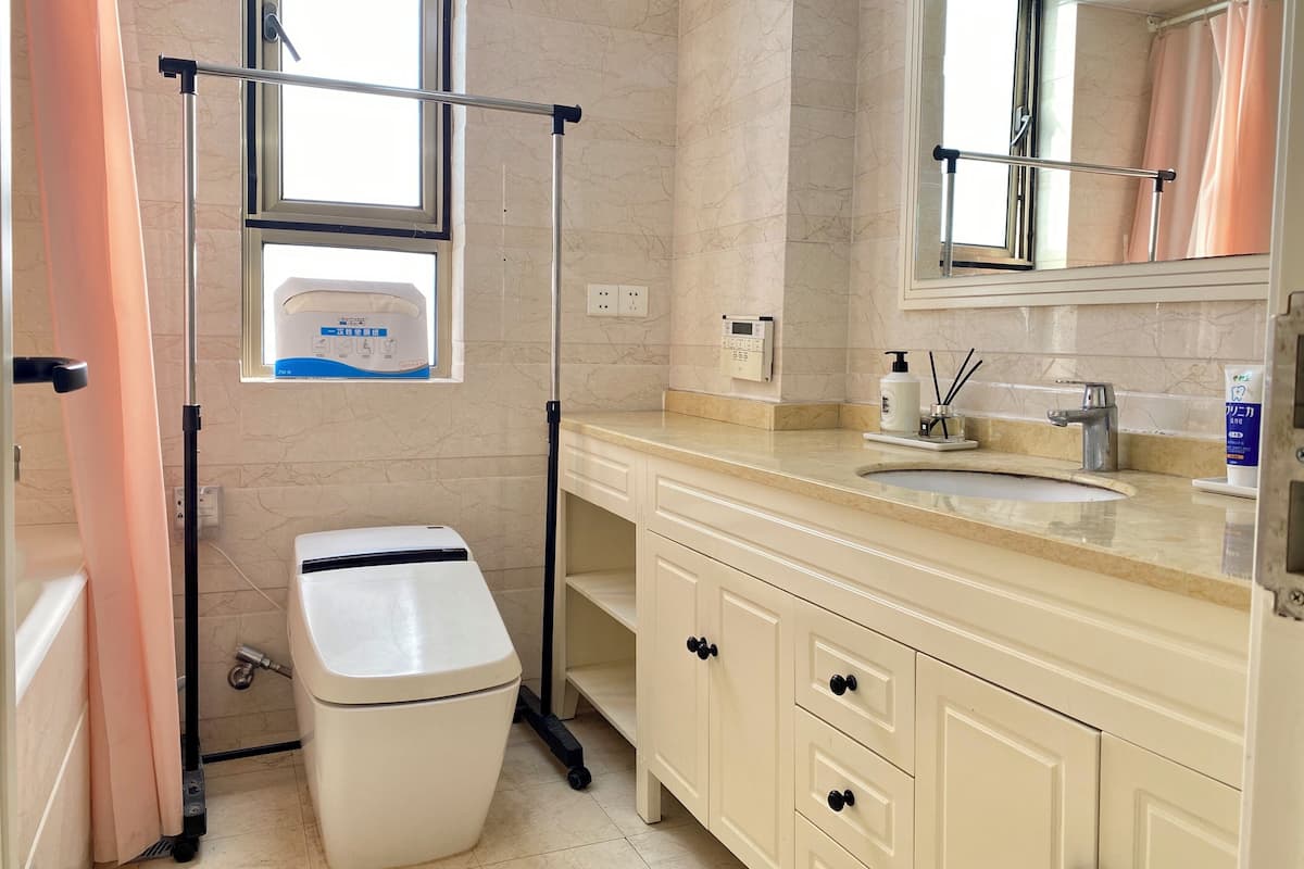 A bathroom with a toilet, a sink, a mirror, a window, and a hand sanitizer dispenser.