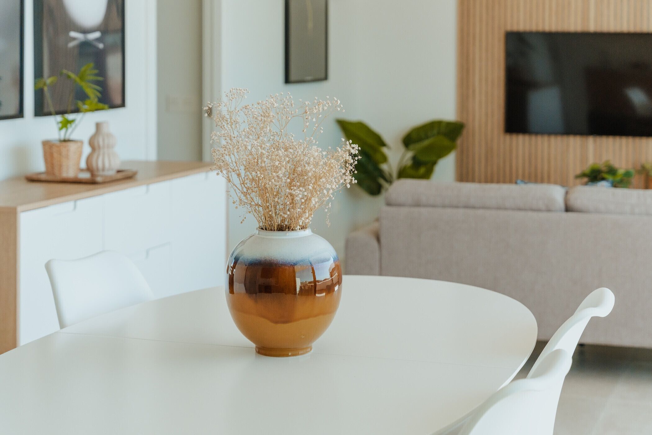Uma sala de estar moderna com uma mesa redonda branca, um vaso com flores secas, um sofá bege e uma televisão de tela plana fixada na parede.