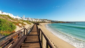 Ein hölzerner Steg führt zu einem Strand, im Hintergrund sind weiße Gebäude zu sehen.