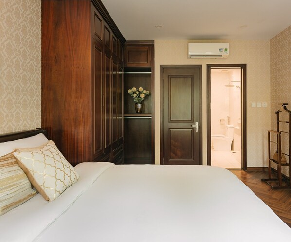 Habitación de hotel con una cama grande, armario de madera, un jarrón con flores, un equipo de aire acondicionado y un baño visible a través de una puerta abierta.