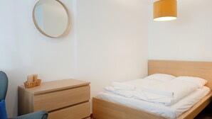 Un lit bien fait, avec du linge de lit blanc, une table de chevet en bois sur laquelle se trouve un panier rempli de bougies, et un miroir rond accroché au mur.