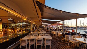 Un espace extérieur aménagé pour les repas, avec des tables et des chaises, donnant sur un port de plaisance où sont amarrés des bateaux.
