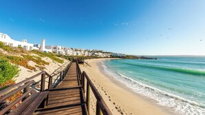 A wooden boardwalk leading to a beach with white buildings in the background. - Luxe Beachside Retreat | Patio | Resort Perks (Langebaan)