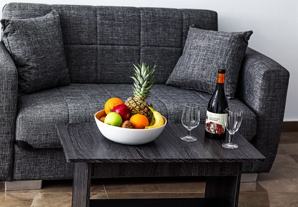 A modern living room with a grey sofa, a wooden coffee table, a bowl of fruit, a bottle of wine, and two wine glasses. - Xaravgi resort suites (Fira)