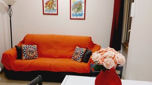 A room with a white table, a red vase with flowers, an orange sofa with patterned cushions, and two framed pictures on the wall. - UN PASSO DAL PORTO (Palermo)