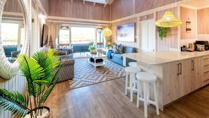 A spacious living area with a large window, a sofa, a coffee table, and a kitchen area with a bar counter and stools. - Luxury Family Chalet | BBQ Deck & Lagoon View (Knysna)