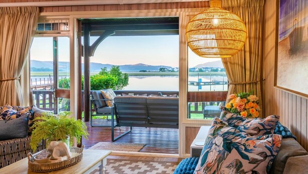 A cozy living room with a view of a lake, a wicker hanging light, and a floral throw pillow. - Luxury Family Chalet | BBQ Deck & Lagoon View (Knysna)
