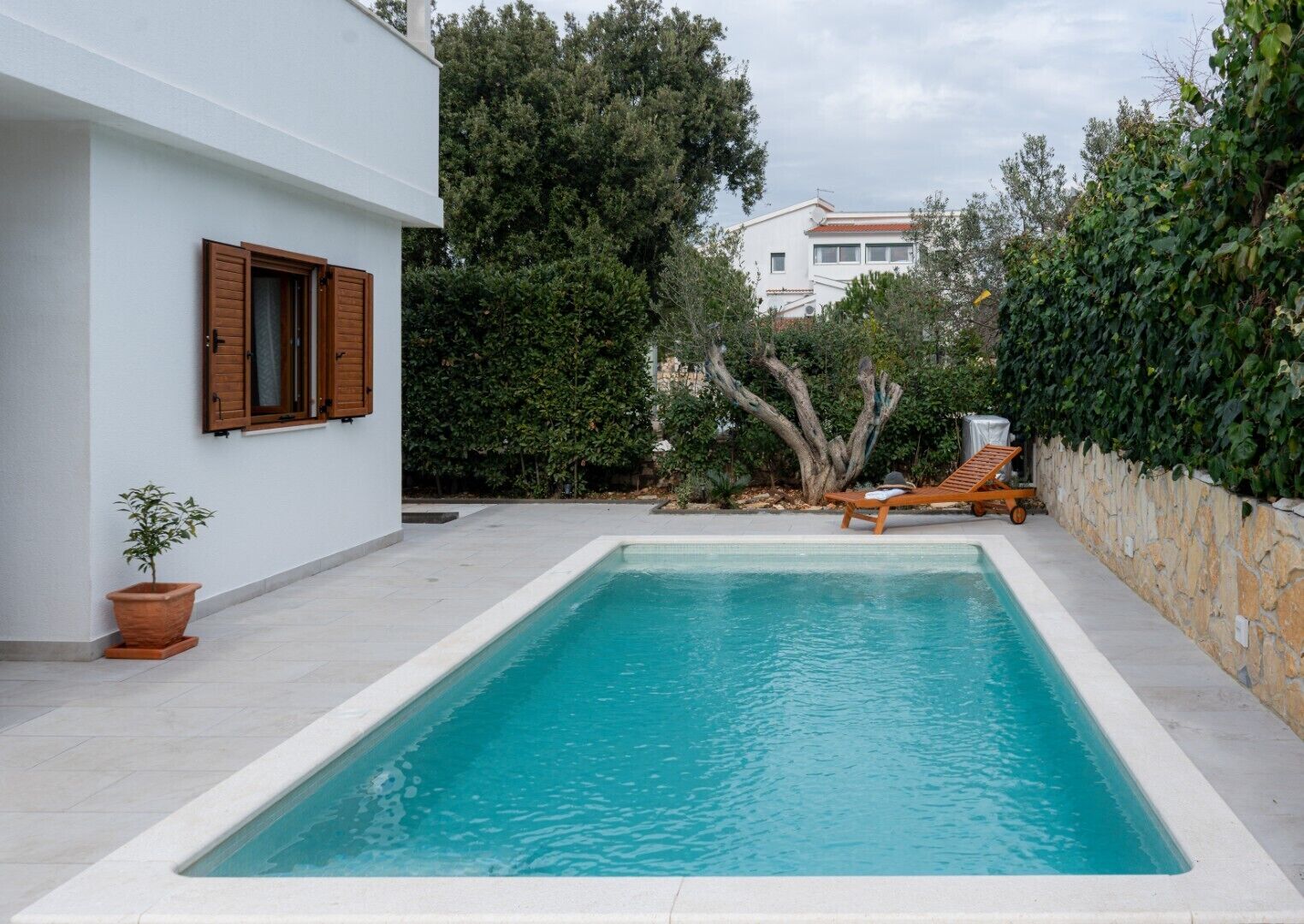Una piscina rettangolare con bordo piastrellato in bianco, una sedia a sdraio in legno e una pianta in vaso.