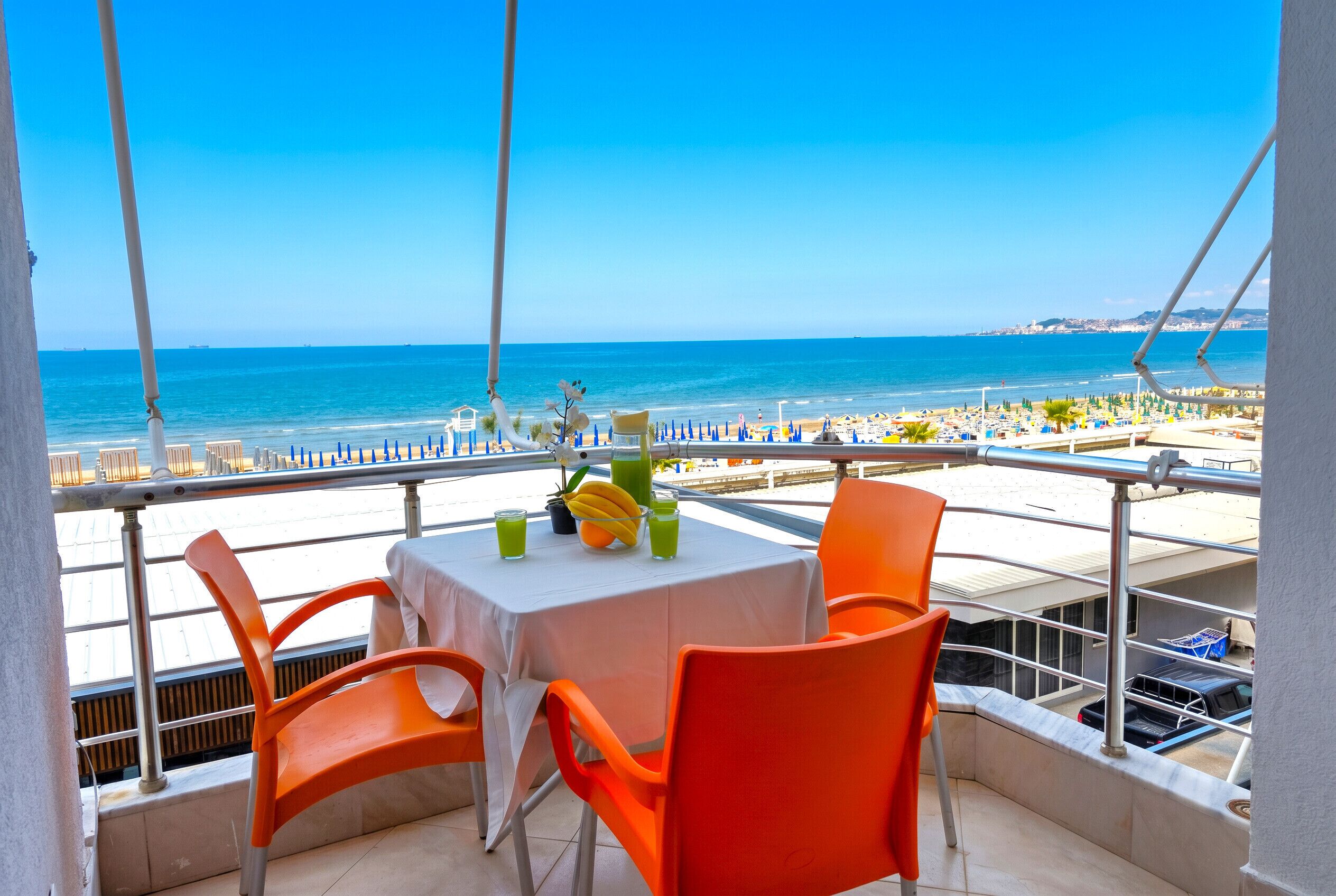 A balcony with a table and chairs, overlooking a beach with umbrellas and a clear sky.