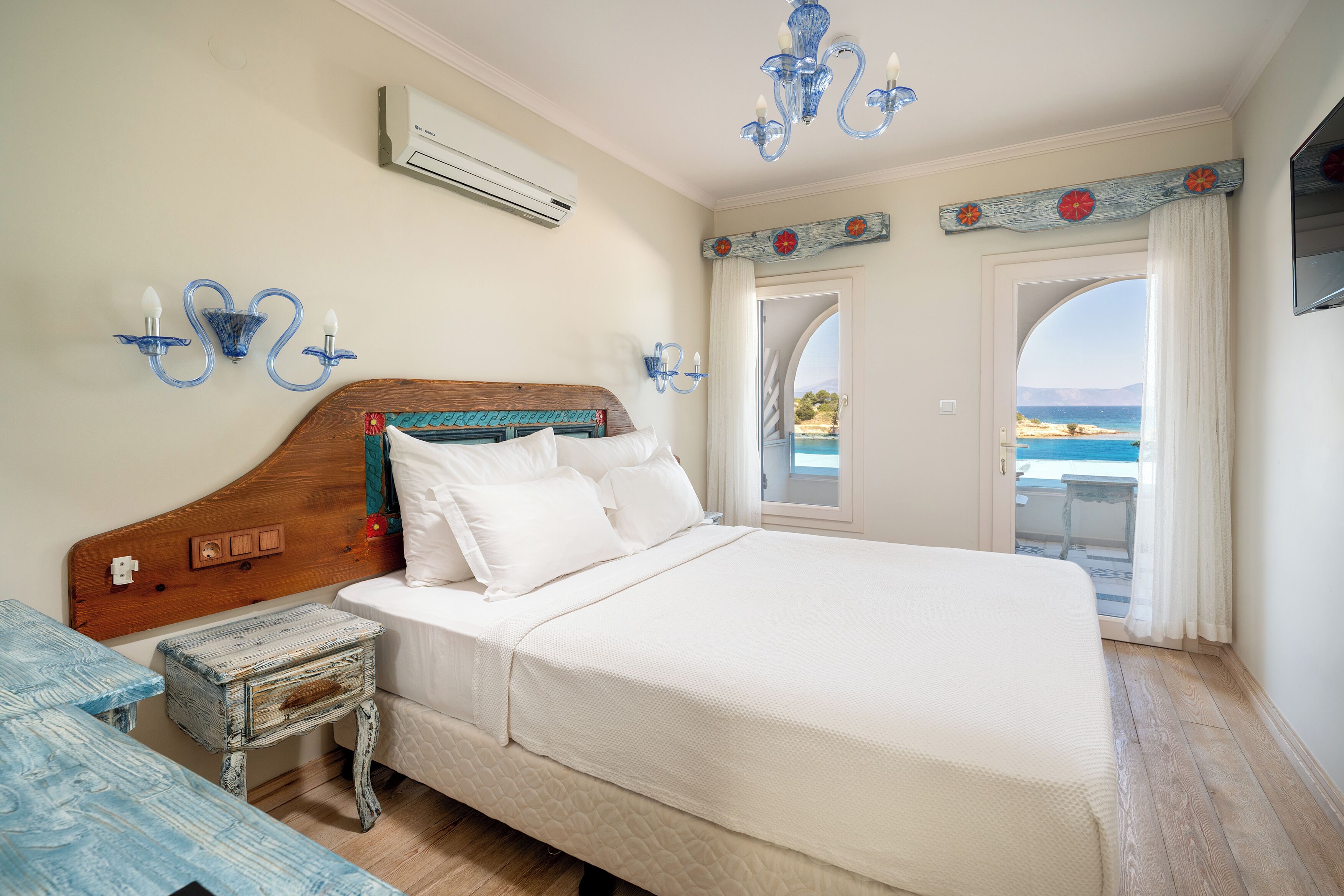 Un dormitorio con cabecera de madera, ropa de cama blanca, una mesita de noche pequeña y vista al mar a través de una puerta abierta.