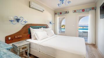 Un dormitorio con cabecera de madera, ropa de cama blanca, una mesita de noche pequeña y vista al mar a través de una puerta abierta.