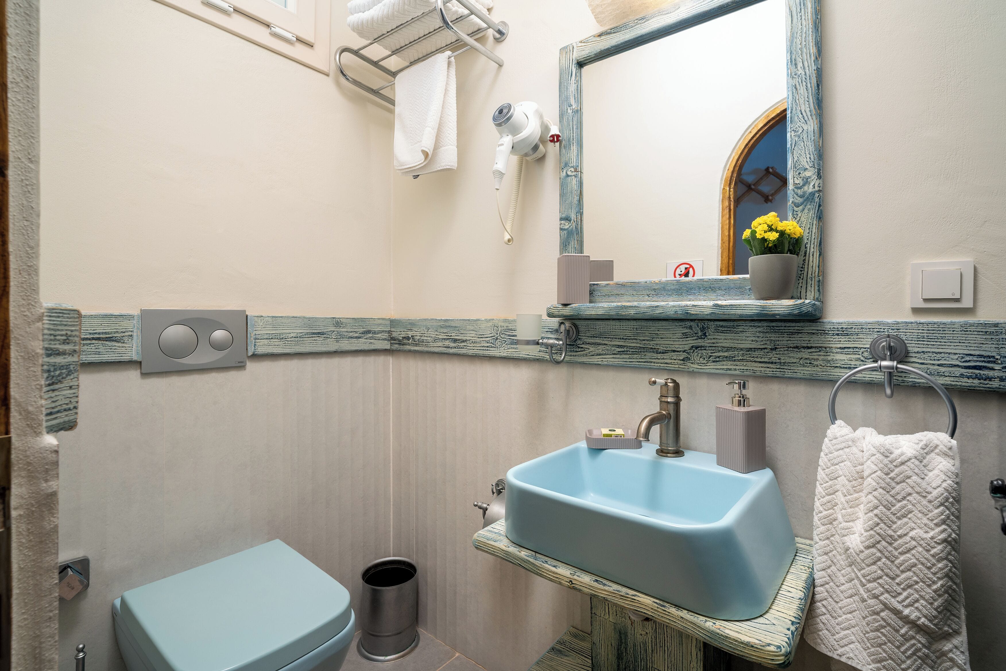 A bathroom with a blue sink, a toilet, a trash can, a towel rack, a mirror, and a hairdryer.