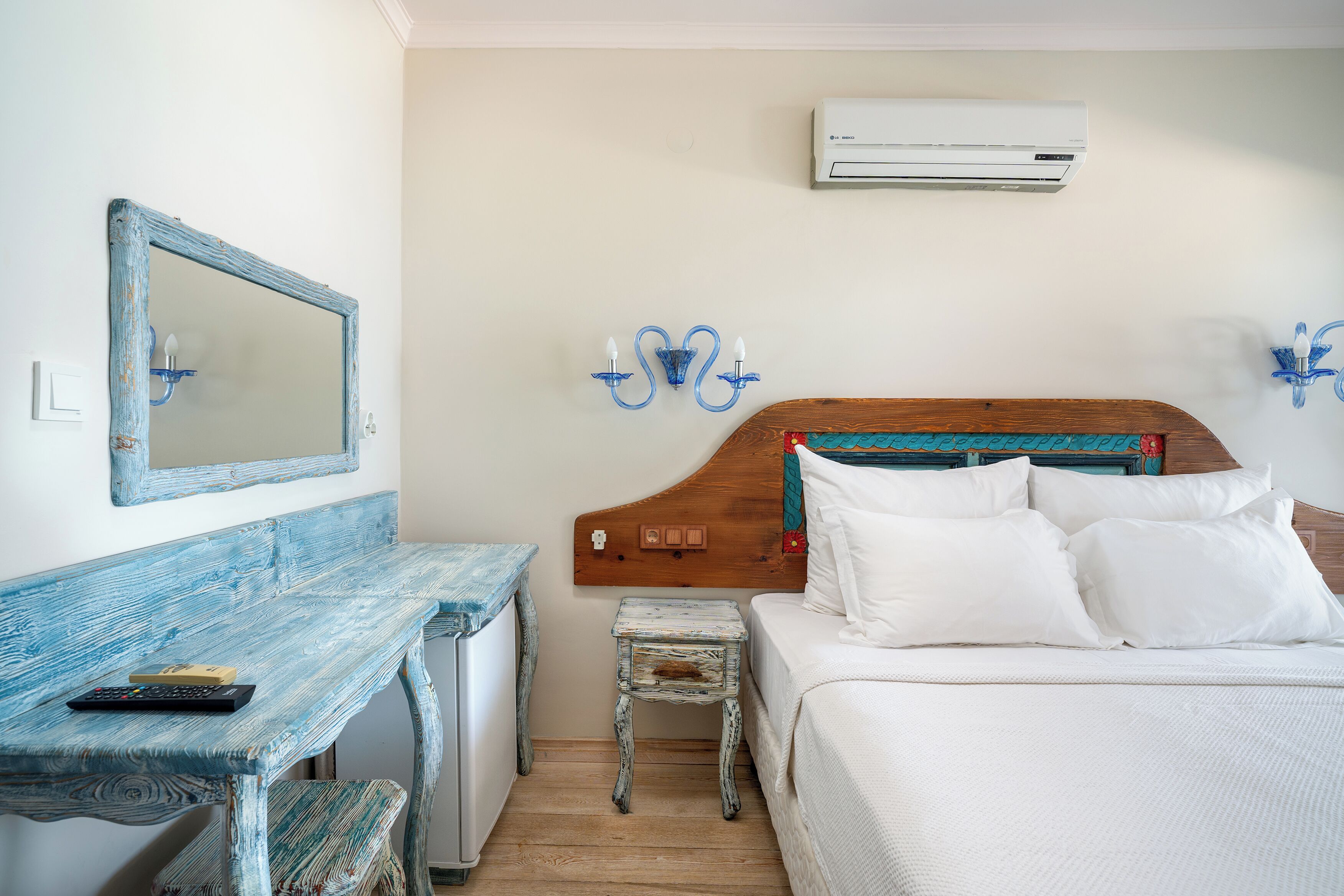 A bedroom with a wooden headboard, white bedding, a small bedside table, a mirror, and an air conditioning unit.