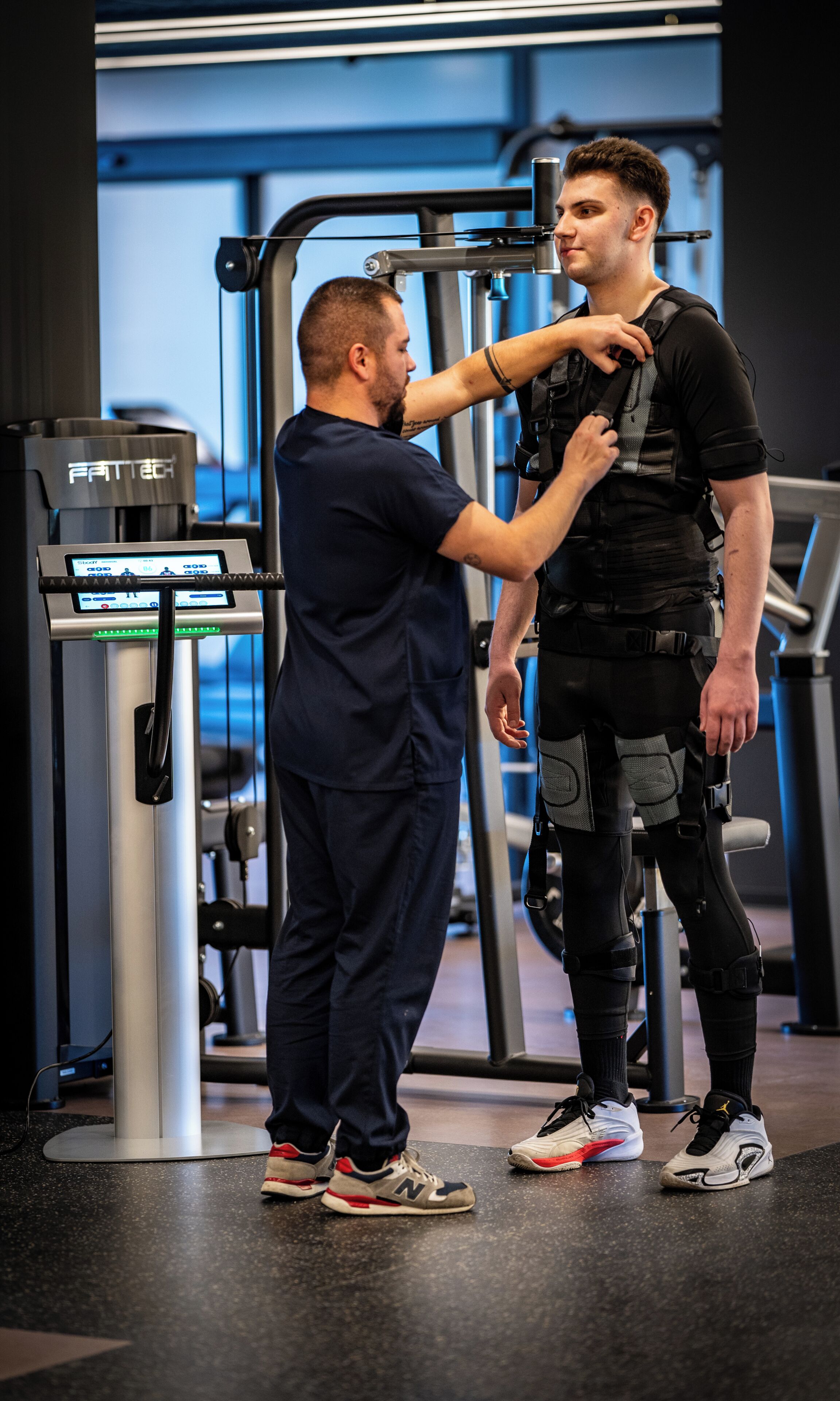 A person being measured by another individual in a gym setting with exercise equipment.