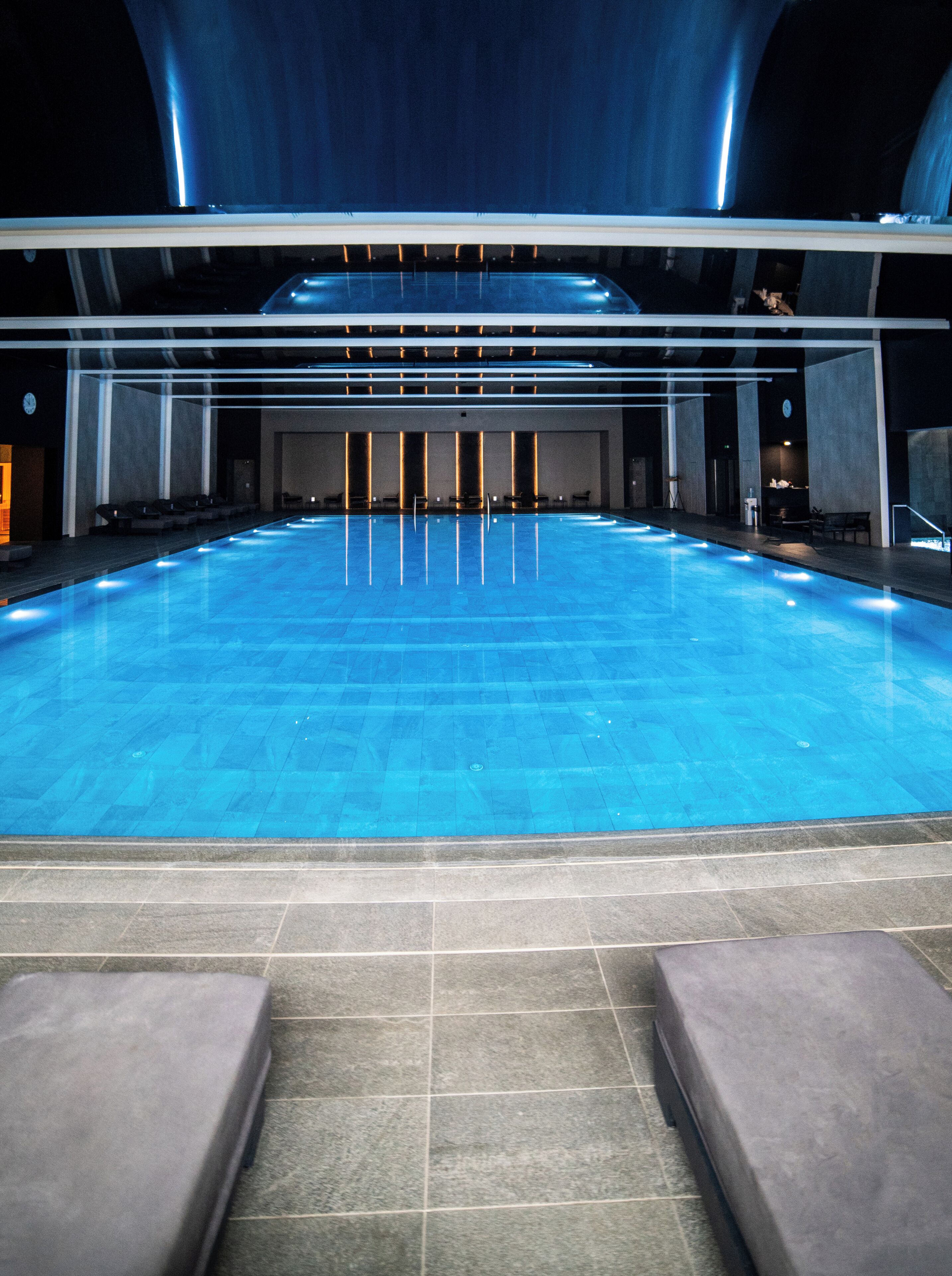 Une grande piscine intérieure avec un sol carrelé, entourée d’espaces aménagés pour s’asseoir et dotée d’un plafond très haut.