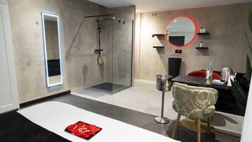 A modern hotel room with a glass-enclosed shower, a bed with a red decorative item, a round mirror, and a dining area with a red chair.