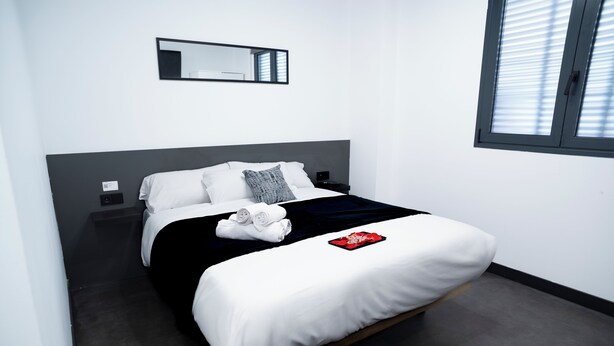 A modern hotel room with a single bed, a bedside table, a nightstand, a mirror, and a window with blinds. - Mad Rooms (Madrid)