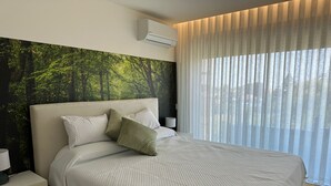 Une chambre moderne avec un grand lit, une tête de lit ornée d’une fresque représentant une forêt, un climatiseur, une table de chevet et des rideaux légers.