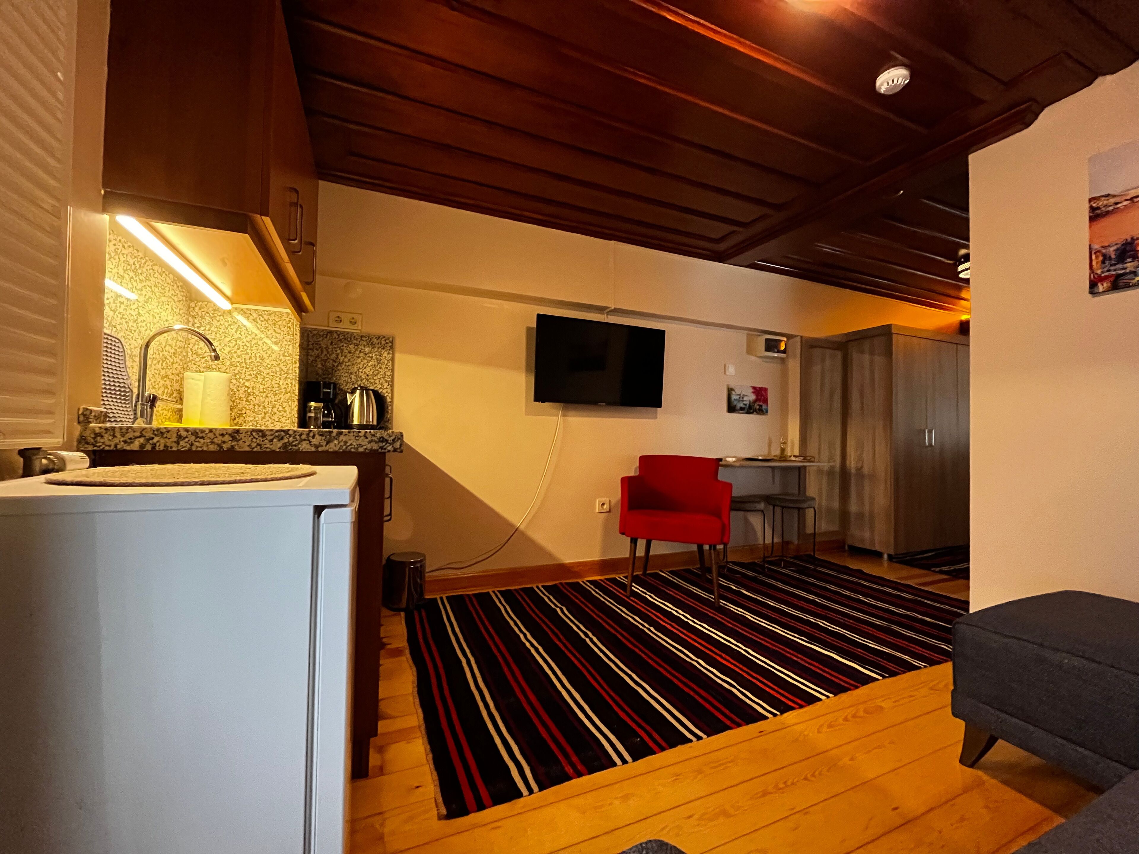 A compact living space with a kitchen area, a red chair, a television mounted on the wall, and a patterned rug.
