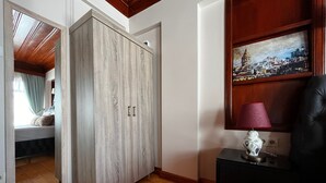 A room with a wooden wardrobe, a bed with a view, a painting, and a lamp on a side table.