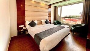 A hotel room with a large bed, a desk, a chair, a ceiling fan, and a window with a view of buildings. - Hotel Roshie Mall Road Mussoorie  (Mussoorie)