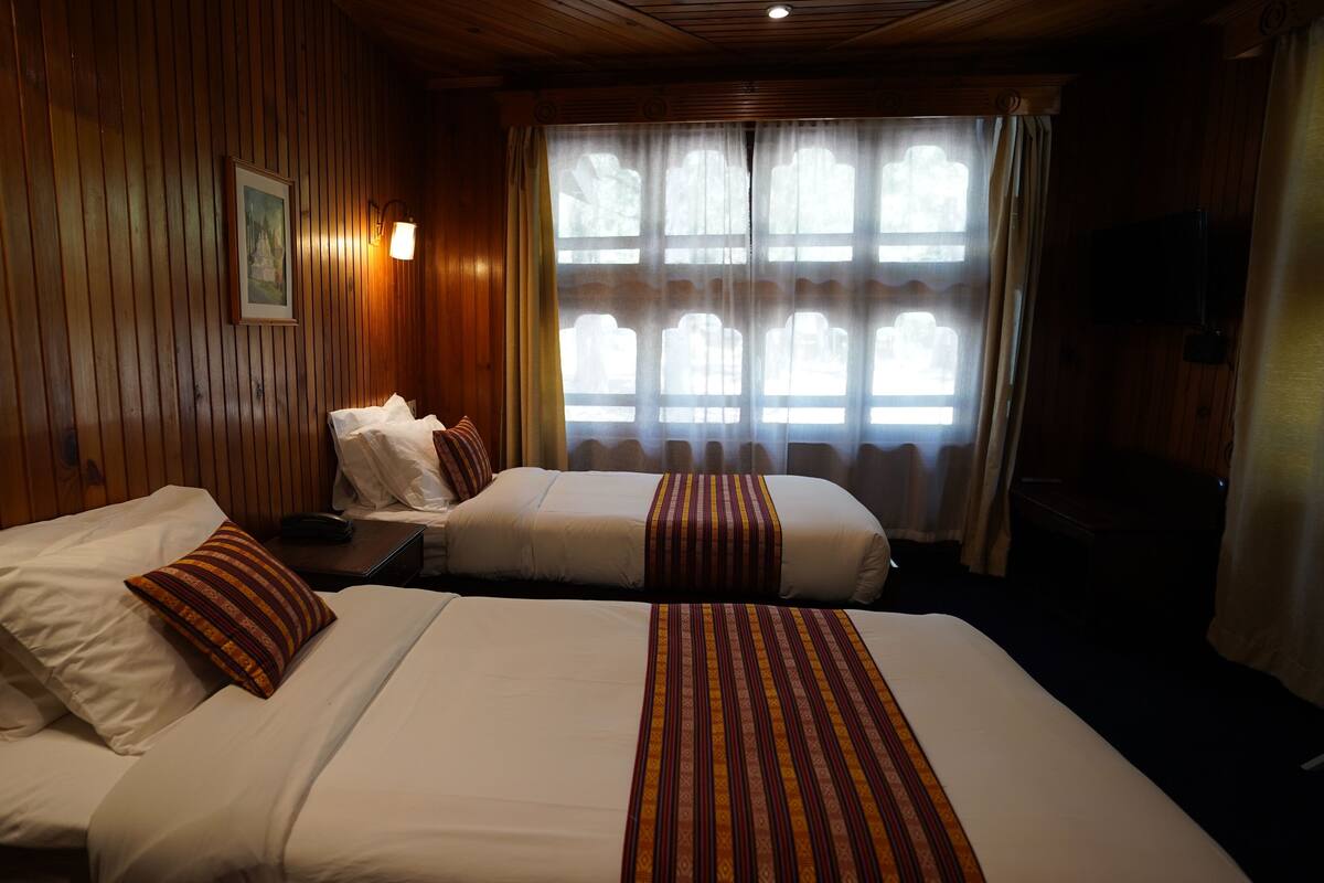 Habitación de hotel con dos camas, paneles de madera, un cuadro en la pared, una ventana con cortinas y un televisor a la derecha.