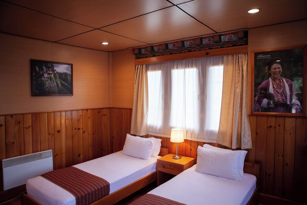 Habitación de hotel con dos camas individuales, paneles de madera, una mesita de noche con lámpara, un cuadro enmarcado en la pared y una ventana con cortinas.