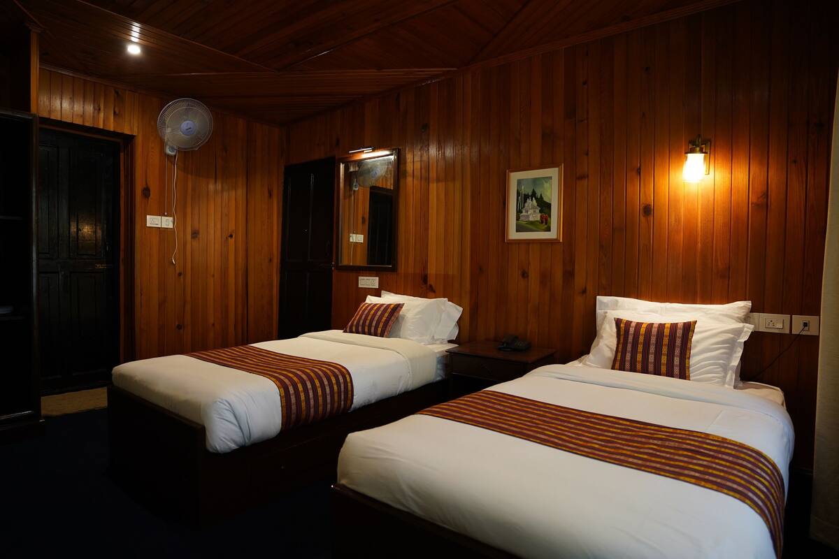 Habitación de hotel con dos camas individuales, paredes de madera, ventilador de techo, un cuadro enmarcado y una mesita de noche con lámpara.