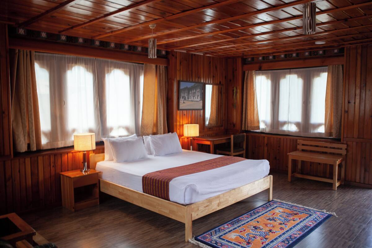 Una habitación con paneles de madera, una cama, dos mesitas de noche con lámparas, un escritorio, una silla y una alfombra.