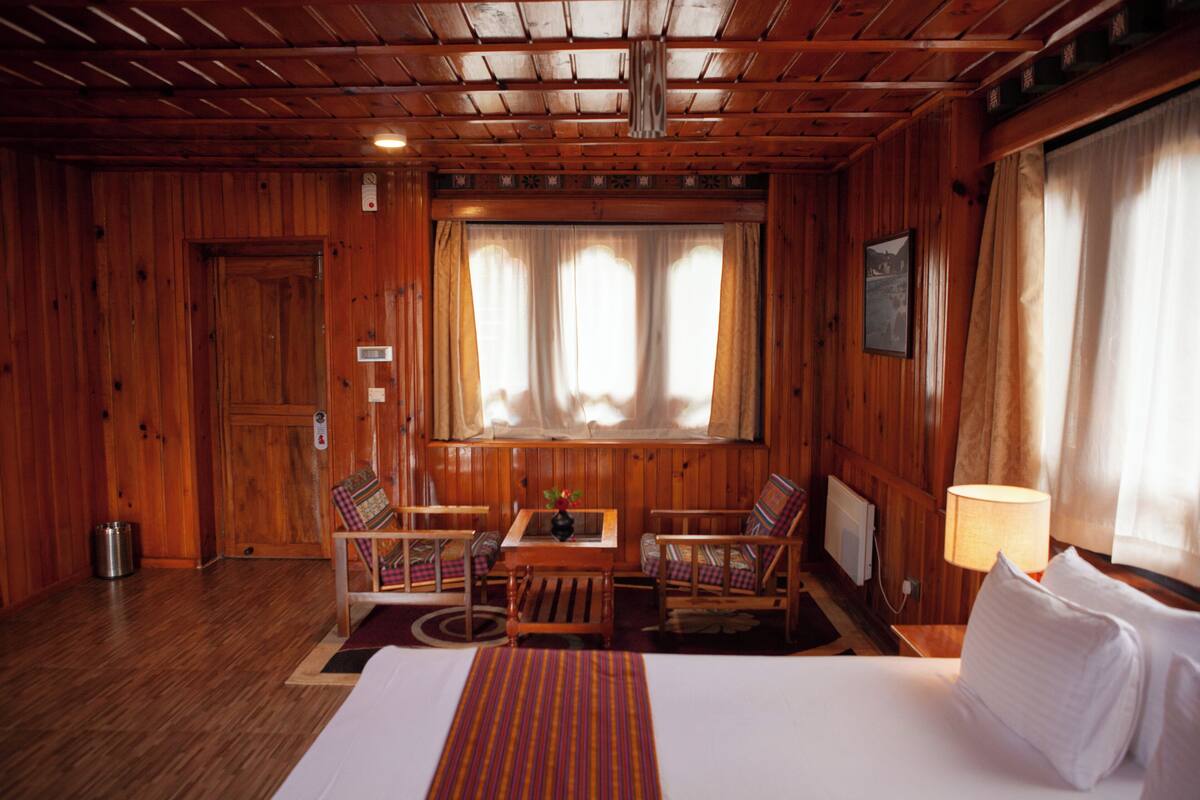 Una habitación tipo cabaña de madera con una cama, dos sillas, una mesita y una ventana con cortinas.