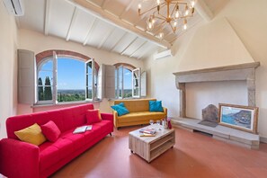 A spacious living room with a red sofa, yellow sofa, and a coffee table. There are large windows with a view, a fireplace, and a chandelier. - Amazing Villa Chimera Green 12 Pool&Gym (Arezzo)