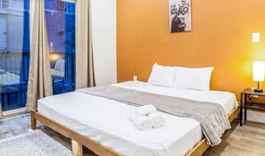 Una camera d'albergo con un letto, una finestra con tende, un quadro appeso al muro e un comodino con una lampada.