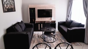 Een moderne woonkamer met een zwarte bank, een zwarte fauteuil, een ronde salontafel en een televisie bevestigd aan een houten kast.