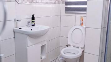 Un baño con lavamanos blanco, espejo y retrete. Hay una ventana con persianas, una zona de ducha y un tapete marrón en el suelo.
