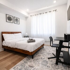 Une chambre moderne avec un lit en bois, un bureau et une chaise. Deux tableaux encadrés ornent le mur, il y a une fenêtre avec des rideaux et une petite table avec une lampe.