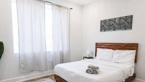 A bedroom with a wooden bed, white bedding, a patterned rug, a nightstand with a lamp, and a geometric wall decoration. - ReeseStreetUnit2inPhilly. (Philadelphia)