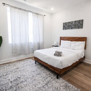 Una camera da letto con un letto di legno, biancheria da letto bianca, un tappeto a motivi, un comodino con una lampada e un decoro geometrico al muro.
