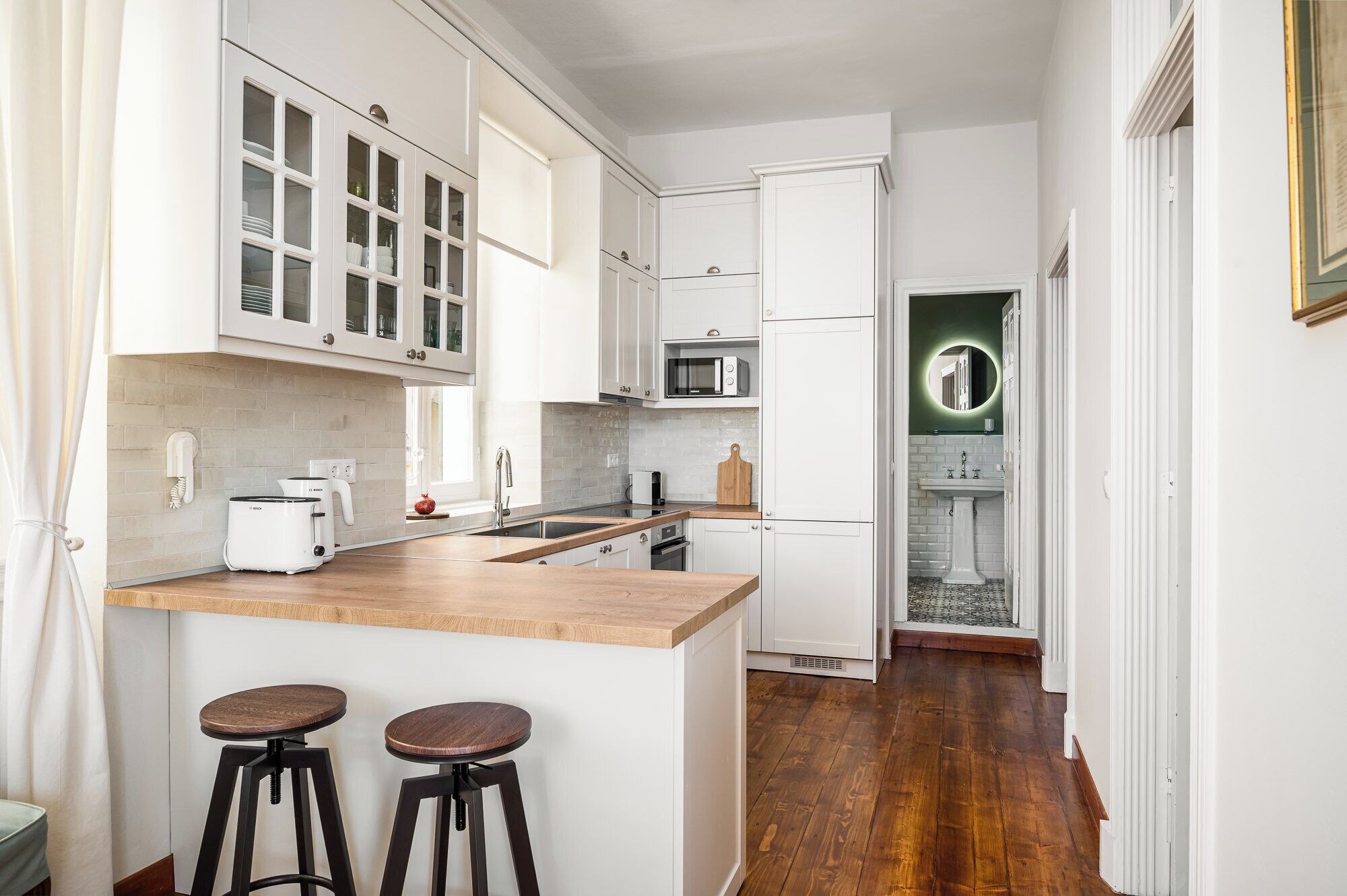 A compact kitchen with white cabinets, a wooden countertop, and two stools.