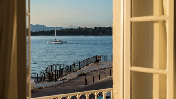 A view of a waterfront with a sailboat, a promenade with a railing, and a distant shoreline with trees and hills. - Casa Milonopoulos by Konnect, Studio & Main House (Corfu)