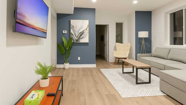 A modern living room with a flat-screen TV, a sofa, a chair, a coffee table, and a potted plant. - Stylish 2BR/2BA near TempleU with Wi-Fi, in-unit laundry & parking (Philadelphia)