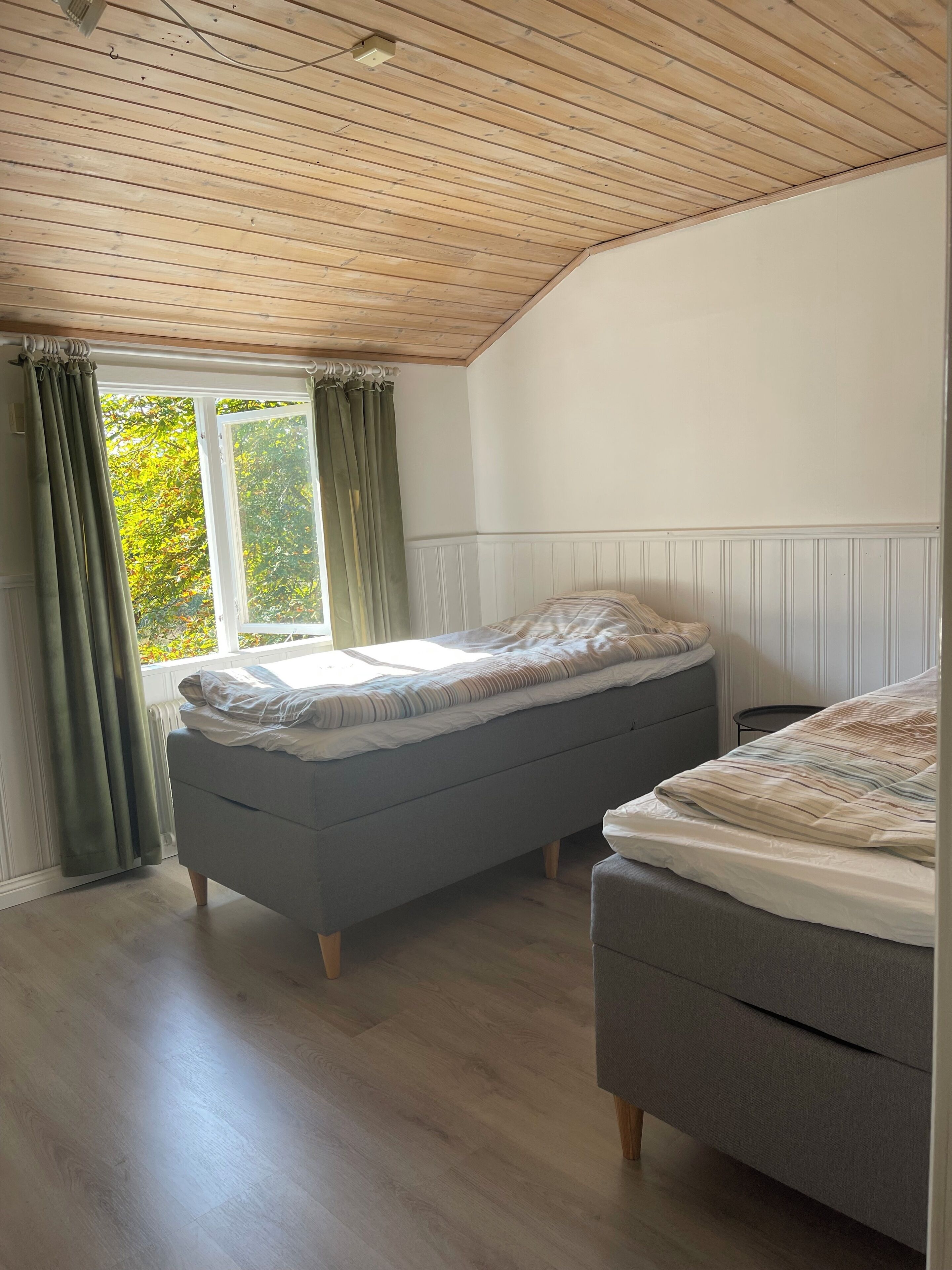 A room with a wooden ceiling, a window with curtains, a bed, and a bench.