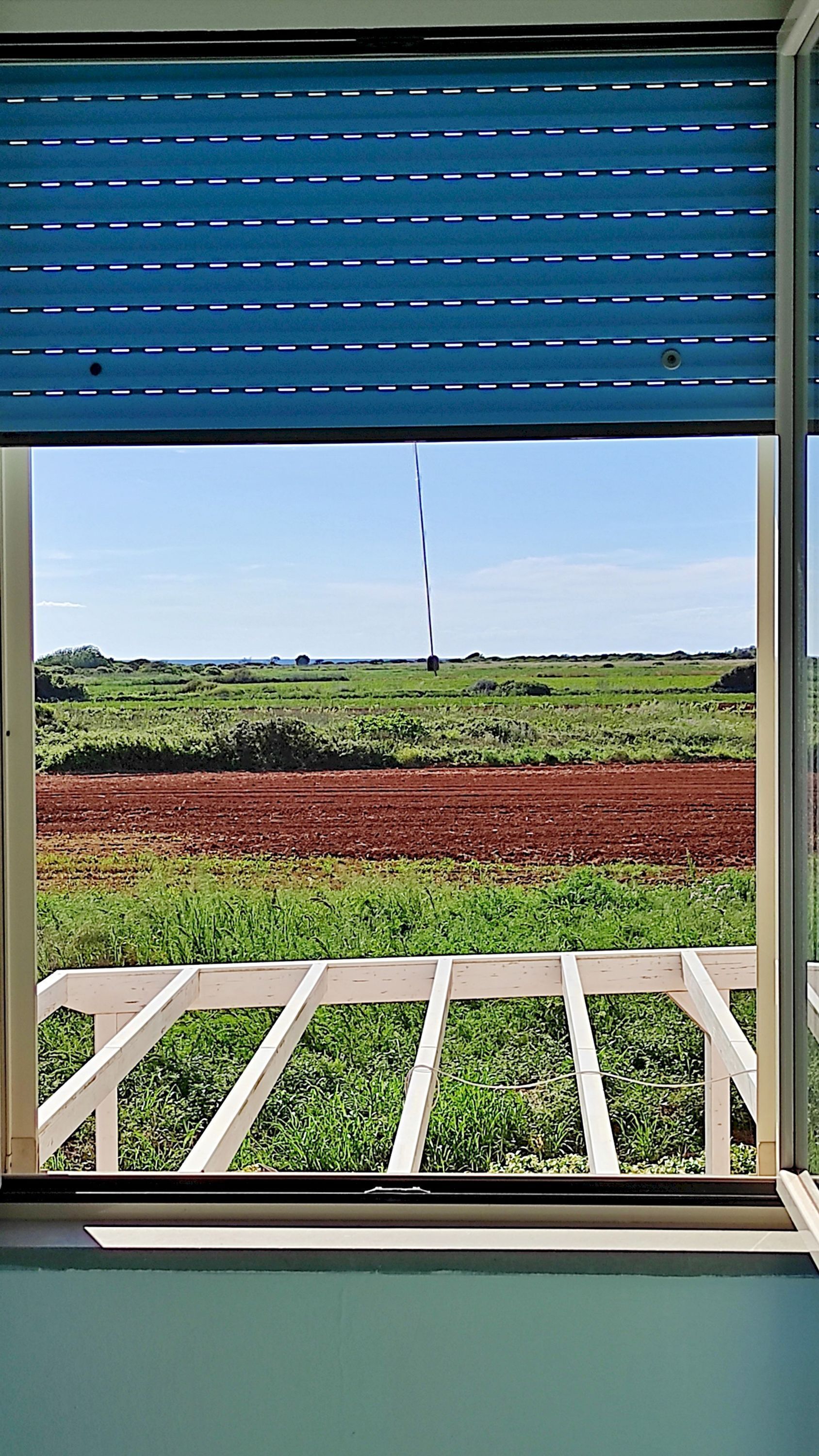 A view through a window with a blue awning, featuring a rural landscape with fields and a clear sky.